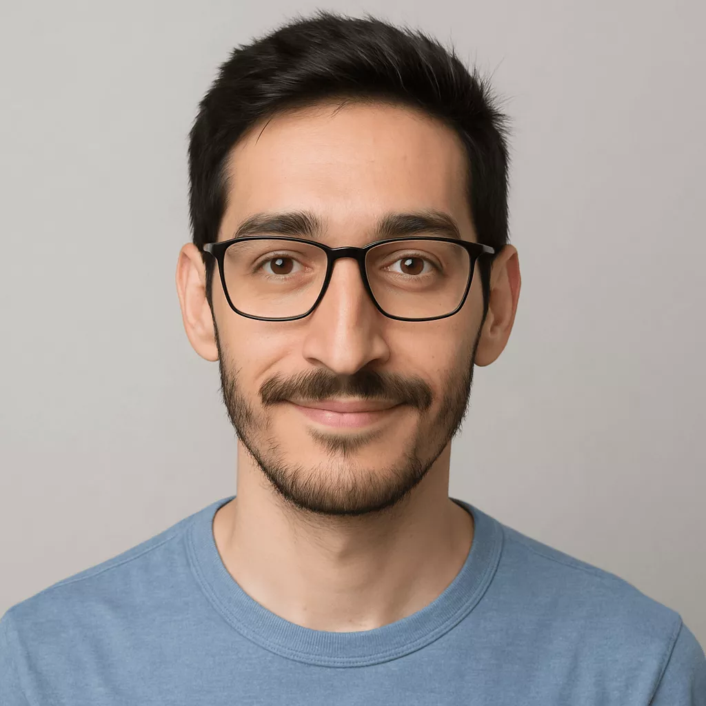 Professional headshot of a young man with short, slightly spiked hair, wearing black rectangular eyeglasses and a muted blue-gray t-shirt, smiling warmly against a neutral light-gray background.